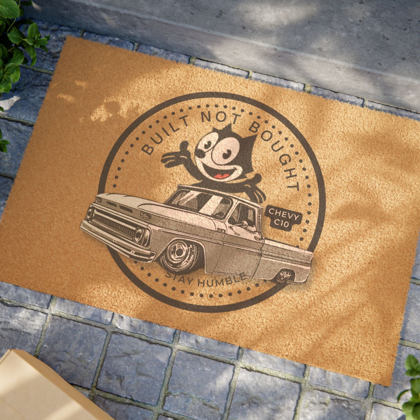 The doormat on a stone paved path outdoors with greenery along the edge.