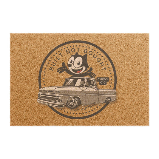 Coir doormat printed with a round Built Not Bought badge: a cartoon cat leaning on a lowered classic pickup truck, with Stay Humble along the bottom.